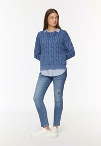Blauwe kabelgebreide trui met verlaagde schouders, gedragen over een lichtblauw en wit gestreept shirt, in combinatie met versleten jeans en witte sneakers.