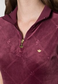 Burgundy zippered polo made of soft, textured fabric with a subtle diamond pattern and gold logo accent. Short collar design.
