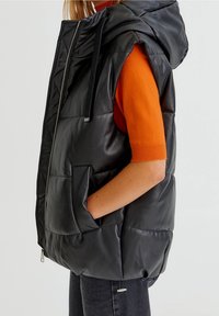 Veste matelassée noire avec capuche, dotée d'une fermeture éclair, de deux poches latérales et d'une texture brillante. Portée par-dessus un haut orange vif.