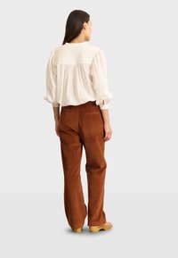 Blusa bianca ampia con yoke testurizzato, pantaloni a gamba larga in velluto a coste marrone e zoccoli di legno, visti da dietro su uno sfondo chiaro.