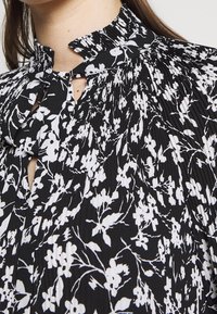 Black and white floral patterned blouse with a pleated high neck, featuring a keyhole opening and tie detail at the collar.