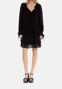 Vestido negro con volantes y mangas largas, con escote en V, que presenta un lazo de cuerda y capas. La modelo lleva zapatos de plataforma gruesa negros.