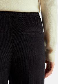 Zwarte corduroy broek met verticale ribbels, voorzien van een achterzak. Gecombineerd met een lichtgekleurde zachte trui, die een textuurcontrast toont.