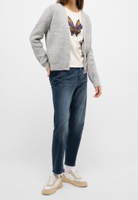 Jasnoszary dziergany kardigan na kremowej koszuli z grafiką motyla, zestawiony z ciemnoniebieskimi dżinsami i kremowymi trampkami z jasnobrązowymi akcentami.