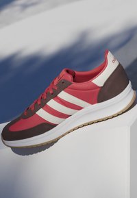 Roter und brauner Adidas-Sneaker mit weißen Streifen und Gummisohle, auf einer weißen Oberfläche mit Schatten im Hintergrund positioniert.