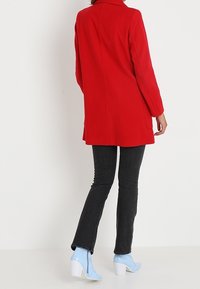 Abrigo rojo con un corte entallado, mangas largas y cuello con solapa. Llevado sobre jeans negros y botas de tacón azul claro.