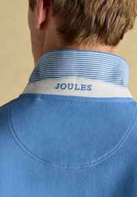 Heled sinine polo särk, millel on tekstuuriga kangas, triibuline krae ja "JOULES" sinise värviga valgel ribal brodeeritud. 