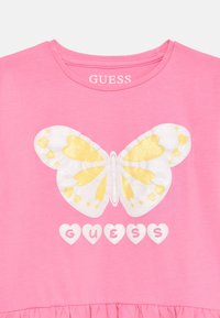 Pink t-shirt featuring a large white and yellow butterfly graphic with heart patterns, and the word "GUESS" in white, heart-shaped letters.