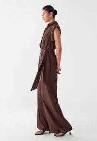 Bruine, mouwloze jumpsuit met een ceintuur in de taille, wijde pijpen en zachte stof. Draag het met zwarte instapsandalen. Model staat in profiel.