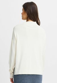 Löstagande pullovertröja i krämfärg, ribbad halsringning, svaga sidoklyftor, mjukt material, långa ärmar och en avslappnad design.