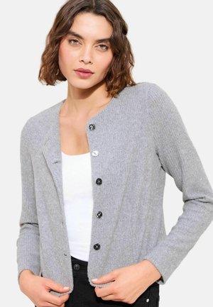 Frau mit kurzem, welligem braunem Haar, die eine hellgraue, mit Knöpfen versehene Strickjacke über einem weißen Top trägt und zur Kamera blickt.