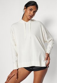 Kvinna som bär en överdimensionerad vit hoodie och svarta shorts, poserar med ena handen vilande på låret mot en neutral bakgrund.