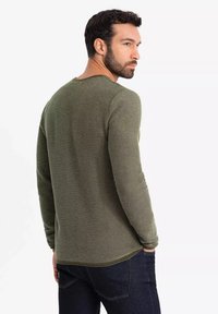 Olivengrünes Langarm-Strickshirt mit einem strukturierten Muster, Rundhalsausschnitt und abgerundetem Saum, getragen mit dunkelblauen Jeans.
