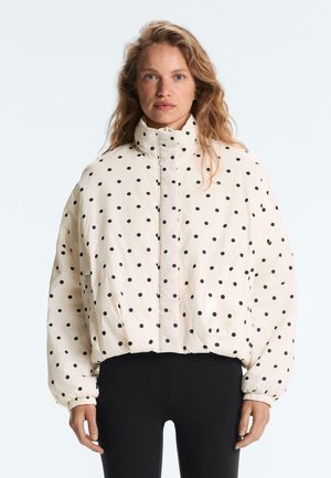 Ženska z valovitimi blond lasmi nosi belo puhasto jakno s črnimi pikami in črne hlače, stoji ob preprostem svetlem ozadju.