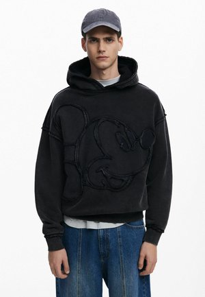 Sort hættetrøje med oversized design, der har et broderet Mickey Mouse ansigt. Ribbestrikkede håndled og kant, båret over en hvid skjorte, med en kasket.