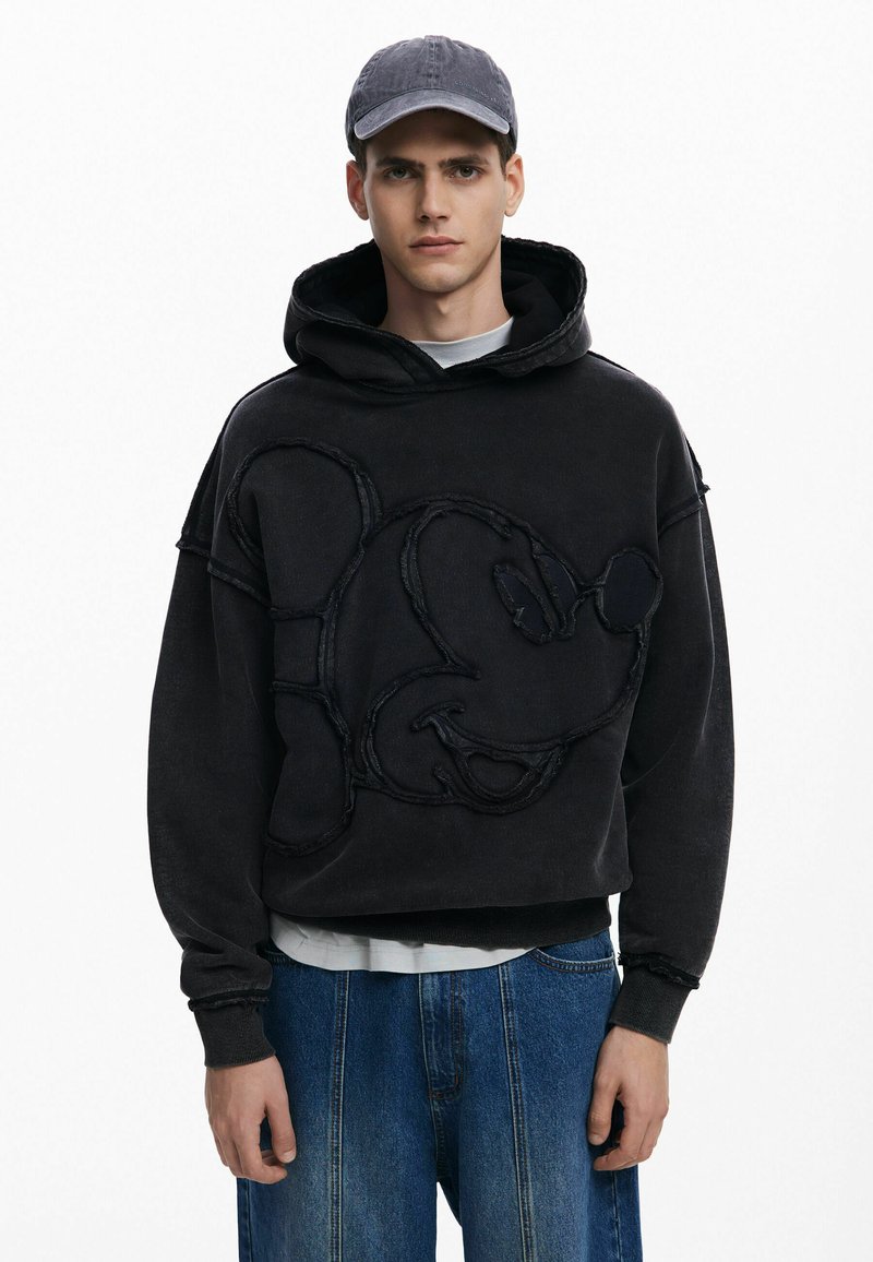 Sort hættetrøje med oversized design, der har et broderet Mickey Mouse ansigt. Ribbestrikkede håndled og kant, båret over en hvid skjorte, med en kasket.