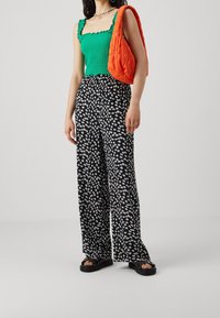 Zielona bluzka na ramiączkach z marszczeniami i falbaniastymi krawędziami, w zestawieniu z czarnymi spodniami o szerokich nogawkach z białym wzorem kwiatowym. Uzupełniona pomarańczową torbą.