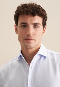Camicia azzurro chiaro con un motivo testurizzato, colletto e bottoni. Il modello è rasato e ha i capelli ricci, su uno sfondo neutro.
