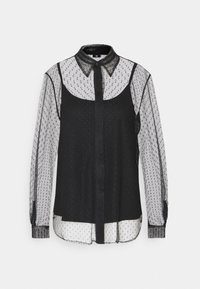 Blouse noire transparente avec motif à pois, devant à boutons, manches longues et une sous-couche blanche contrastante. Le col est en tissu en maille noire.
