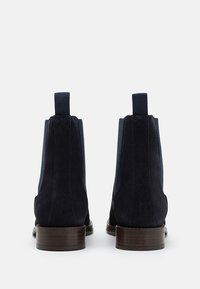 Paire de bottines en daim noir avec languettes de traction et talons en bois empilés vues de dos sur un fond blanc.