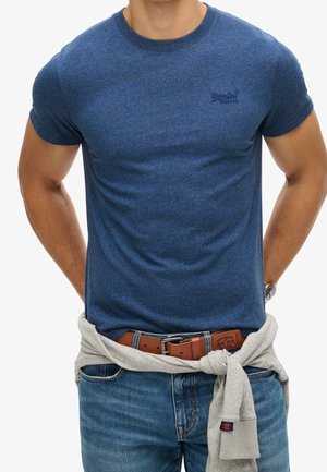 Mężczyzna w niebieskiej koszulce z krótkim rękawem, z szarym swetrem przewiązanym w talii oraz niebieskich dżinsach z brązowym skórzanym paskiem.