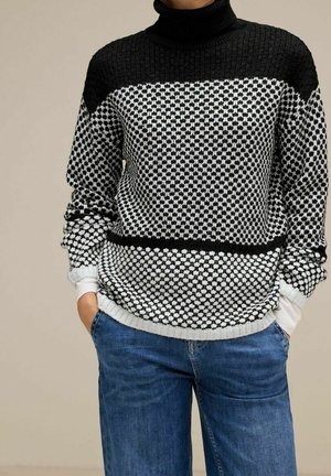 Henkilö pukeutuneena mustavalkoiseen, kuvioituun pooloneuleeseen, kädet sinisten farkkujen taskuissa, beige tausta.