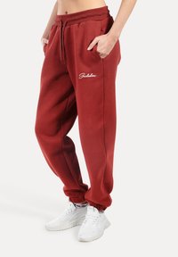 Czerwone spodnie dresowe z regulowanym paskiem w talii, bocznymi kieszeniami i białym logo na udzie. Ściągacze na kostkach. Noszone z białymi trampkami.