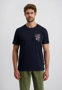 Man in een marineblauwe grafische T-shirt met de tekst "Pierre Cardin" en abstracte vormen, gecombineerd met groene corduroy broek, staande tegen een effen achtergrond.