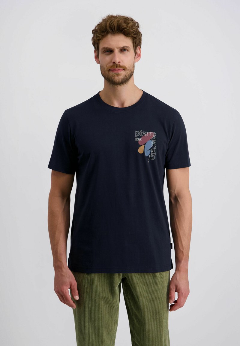 Man in een marineblauwe grafische T-shirt met de tekst "Pierre Cardin" en abstracte vormen, gecombineerd met groene corduroy broek, staande tegen een effen achtergrond.