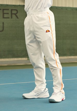 Osoba ubrana w białe sportowe spodnie z pomarańczowymi paskami po bokach i białe buty sportowe stojąca na niebieskim krytym boisku.