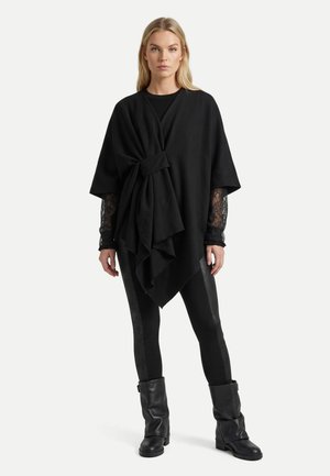 Femme portant un poncho noir style cache-cœur avec un haut à manches en dentelle, un pantalon noir moulant avec des bandes latérales et des bottes noires mi-mollet, debout droite.