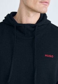 Mann iført en svart teksturert hettegenser med "HUGO" brodert i rødt på brystet, som viser et delvis sidebilde av ansiktet og halsen.