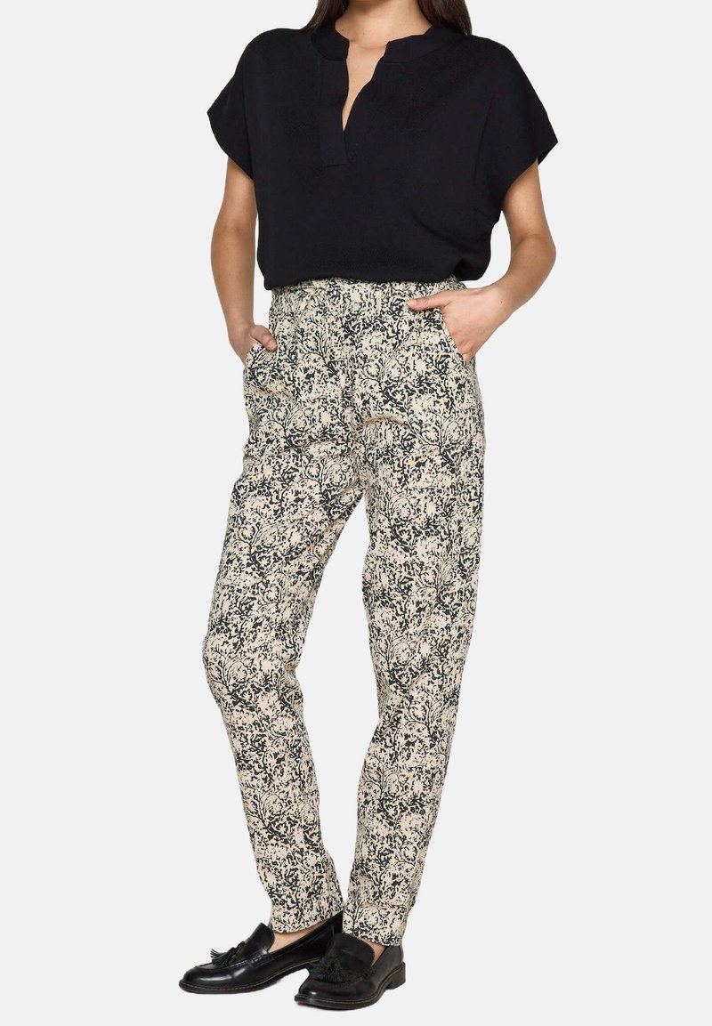 Zwart T-shirt met korte mouwen, losse pasvorm; gemusterde crèmekleurige broek met zwart bloemenpatroon; platte zwarte schoenen; zakken op de broek.