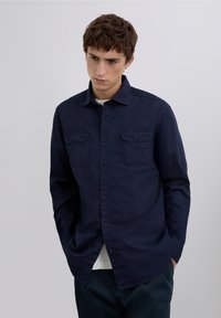 Marineblaue Hemd mit Knopfleiste aus einem glatten Baumwollmischgewebe, ausgestattet mit zwei Brusttaschen und einem klassischen Kragen, getragen über einem hellen Hemd.