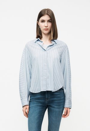 Νεαρή γυναίκα που φοράει ανοιχτό μπλε ριγέ πουκάμισο και μπλε τζιν, στέκεται μπροστά σε απλό λευκό φόντο.