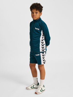 Dreng står med hænderne bag ryggen iført teal lynlåstrøje og shorts med hvide og sorte chevronstriber og hvide sneakers.