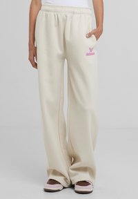 Pantaloni della tuta color crema con vita elastica, caratterizzati da un logo ricamato rosa sul lato sinistro e gambe ampie e fluide.