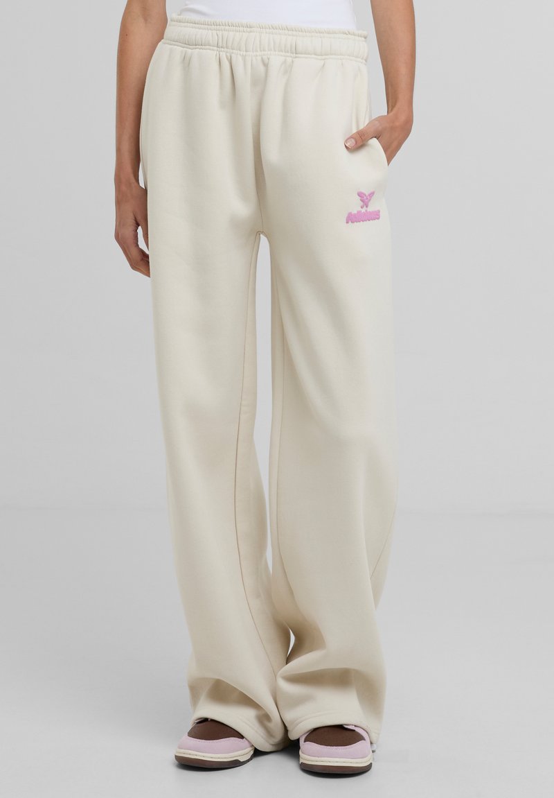 Pantaloni della tuta color crema con vita elastica, caratterizzati da un logo ricamato rosa sul lato sinistro e gambe ampie e fluide.