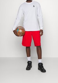 Weißes Langarmshirt mit schwarzem Logo, rote Shorts, schwarze High-Top-Sneaker und einen braunen Basketball mit weißem Logo in der Hand.