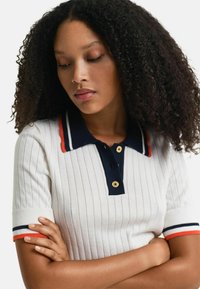 Camisa polo blanca con cuello azul marino y acentos a rayas en naranja y azul. Presenta botones dorados y mangas cortas.
