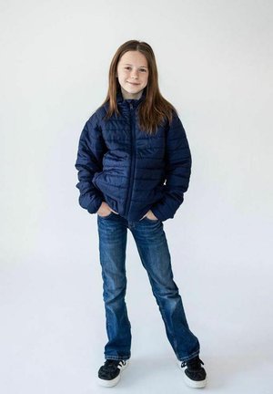 Tyttö seisoo kädet taskuissa, yllään tummansininen toppatakki, siniset farkut ja mustat tennarit vaalean yksivärisen taustan edessä.