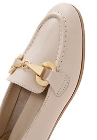 Mocassino in pelle beige con punta arrotondata, dettagli cuciti e un accento decorativo in metallo dorato sulla tomaia. Texture liscia.