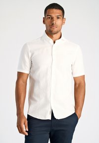 Man in een aansluitend, kort mouw wit overhemd en donkere broek, staand met één hand in zijn zak tegen een effen achtergrond.