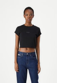 Melnā īsā T-krekla ar īsām piedurknēm un apaļu kakla izgriezumu, uz kuras krūšu daļas ir neliela Calvin Klein logo. Saskaņots ar tumši zilām džinsēm.