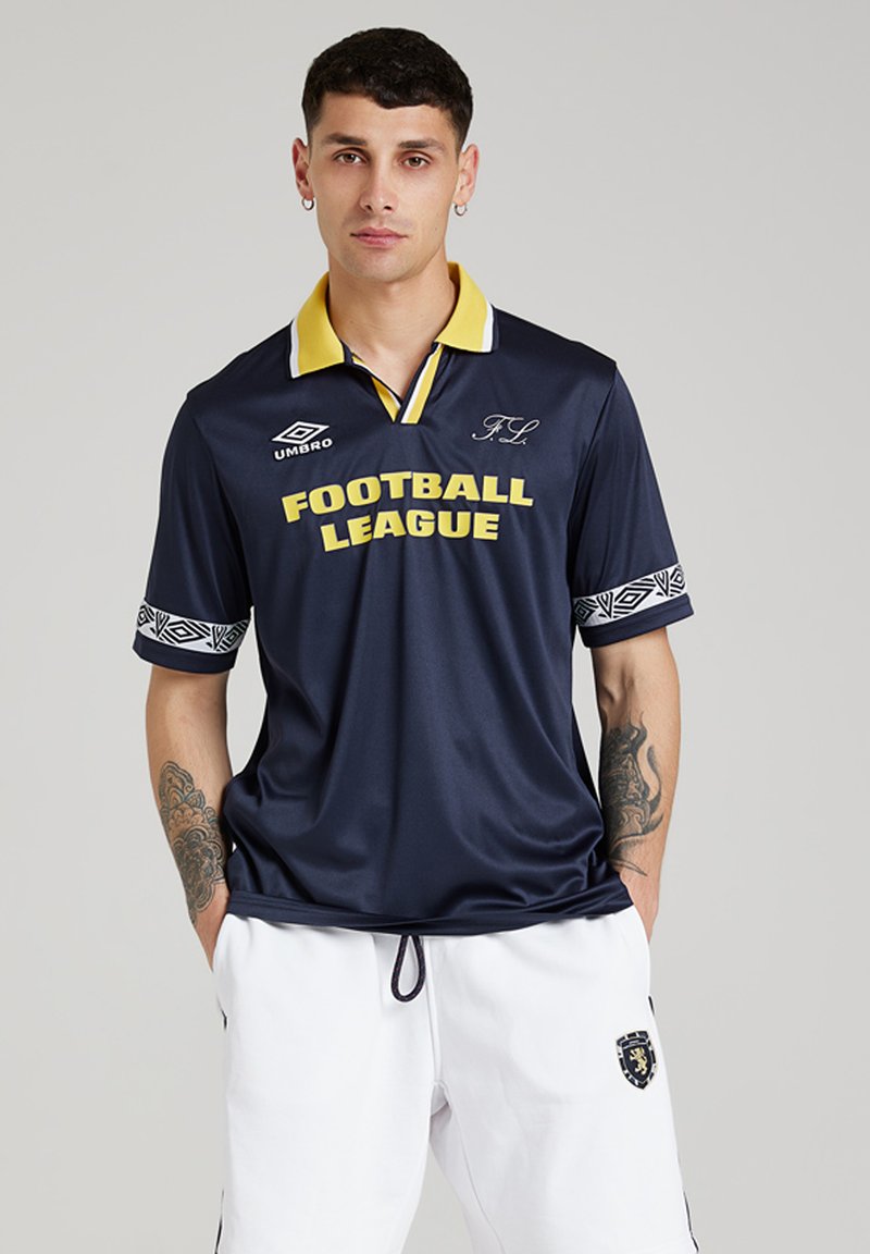 Tânăr cu cercei și tatuaje, îmbrăcat cu un tricou al "Ligii de Fotbal" de culoare bleumarin, cu guler galben și pantaloni scurți albi cu un blazon.