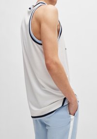 Débardeur de sport blanc avec des bordures bleu marine et bleu clair, présentant une texture en maille et une coupe décontractée. La main du modèle est dans sa poche.