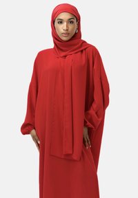 Robe rouge fluide à manches longues, avec un hijab assorti. Tissu lisse avec peu de détails, créant une silhouette élégante et harmonieuse.