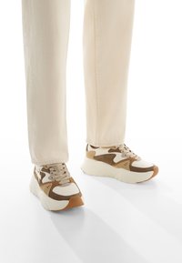 Sneakers beige con accenti marroni e bianchi, materiali in suede e mesh, suola spessa e punta rotonda. Indossati con pantaloni tapered beige chiaro.