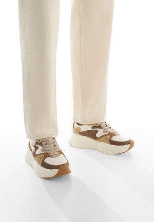 Baskets beiges avec des accents brun et blanc, en matériaux en daim et en maille, semelle épaisse et bout rond. Portées avec un pantalon tapered beige clair.