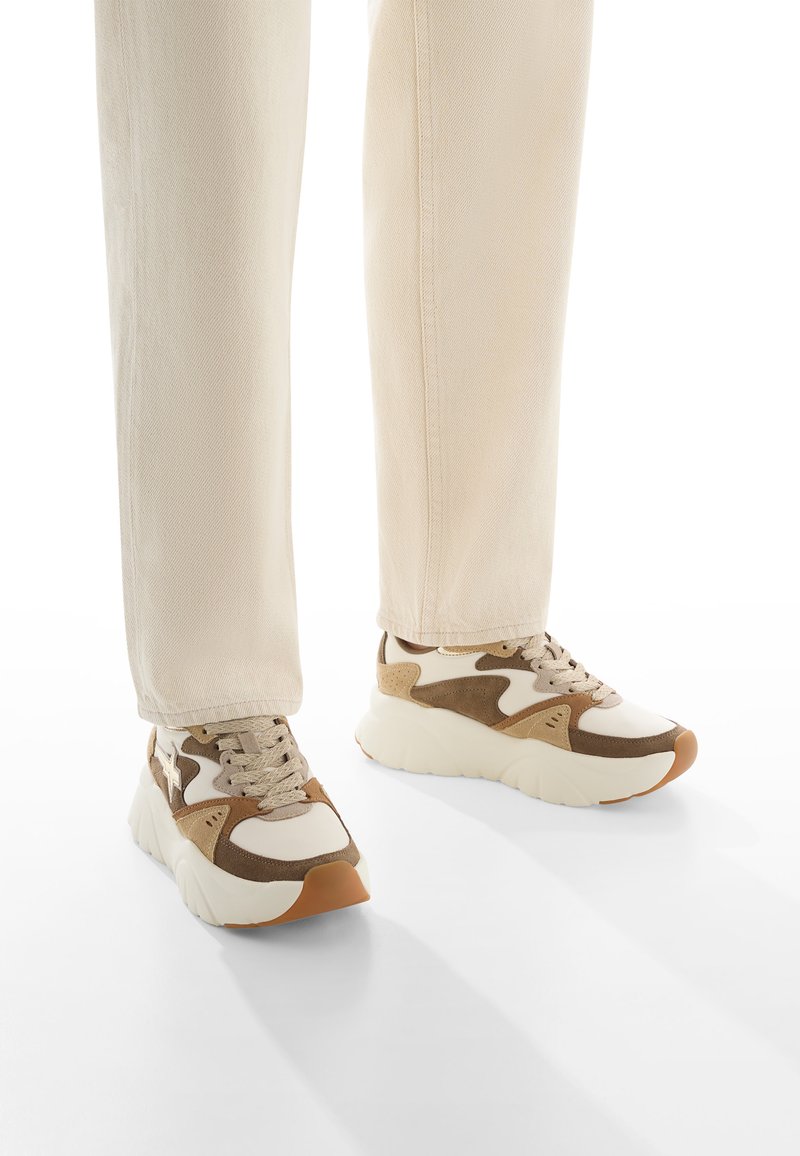 Sneakers beige con accenti marroni e bianchi, materiali in suede e mesh, suola spessa e punta rotonda. Indossati con pantaloni tapered beige chiaro.